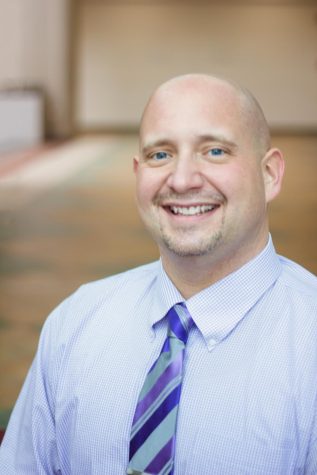 A professional headshot of Zach Clark. Clark is a middle aged white man wearing a light blue shirt and a blue tie, smiling at the camera.