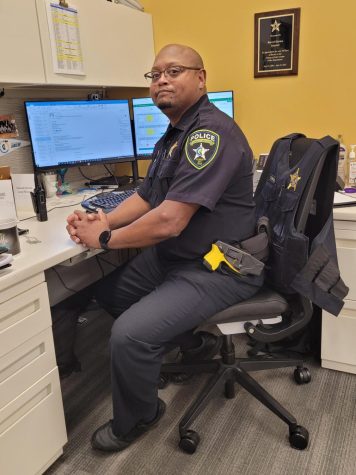 Lt. Harlan, sitting in his desk in his police uniform. He has his hands folded in front of him casually and is smiling lightly at the camera.