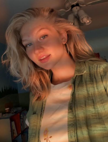 A selfie of Allie Jones. Jones is a young woman with wavy platinum blonde hair that reaches past her shoulders. She is wearing a green flannel over a white shirt and is lightly smiling at the camera.