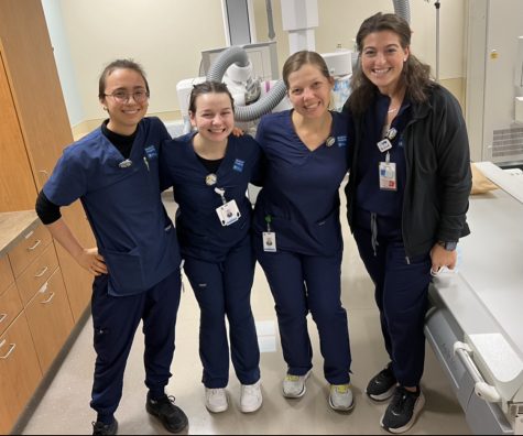 Four people with their arms around each other smiling for the camera. They are all dress in navy blue scrubs and stand in an xray room.