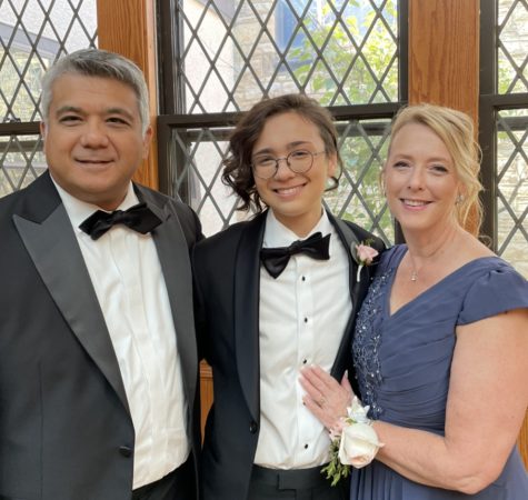 A picture of Castor with their parents in formal attire. Them and their father are both in suits. Their mother is in a dark blue dress and has a corsage on her wrist. Castor is between their parents. Everyone is smiling.